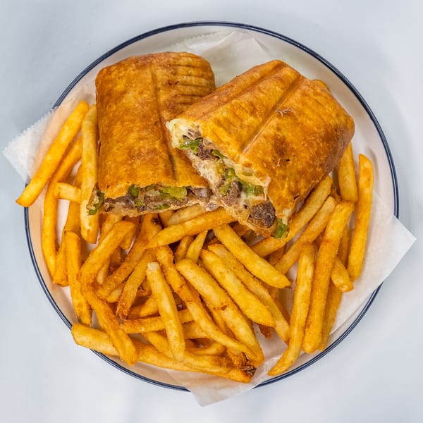 A golden-brown Philly cheesesteak sandwich, cut in half, served with a side of crispy french fries.